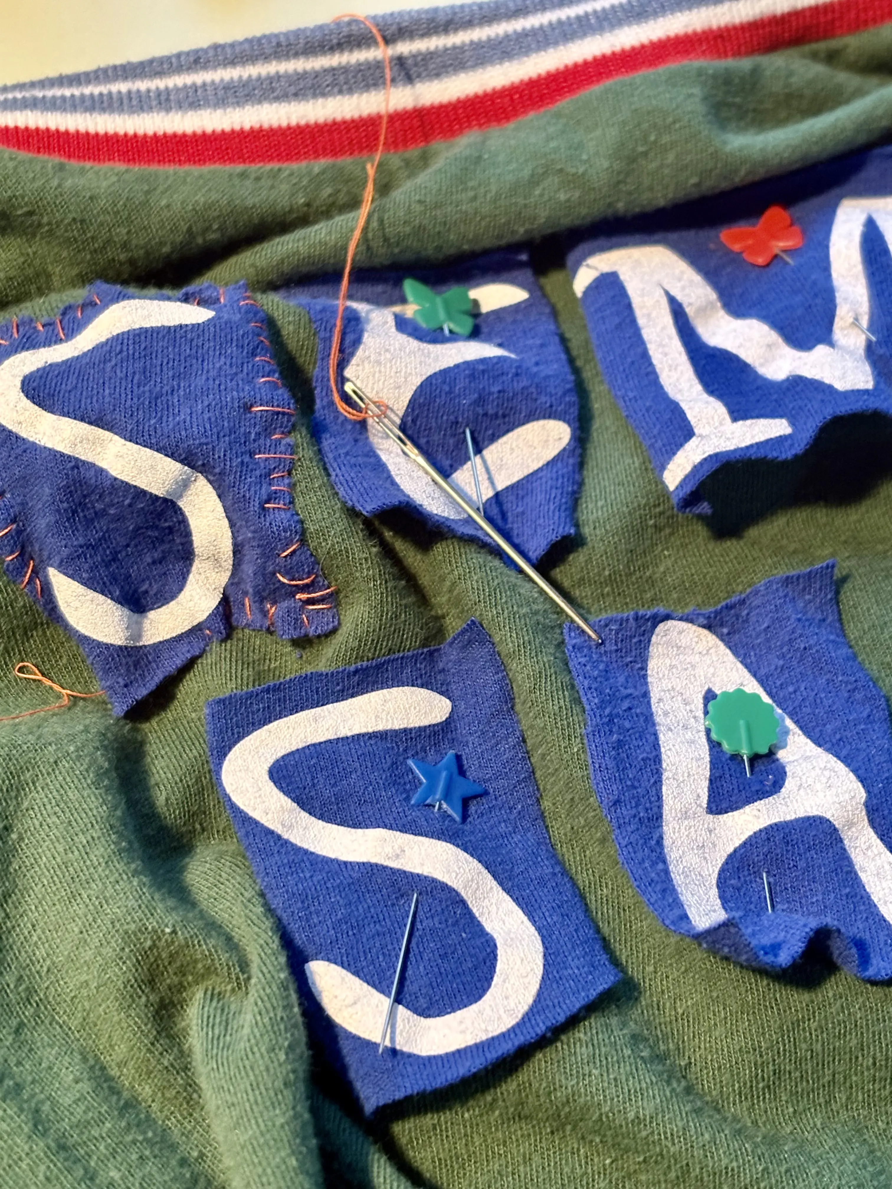 Cut blue fabric lettering being hand-stitched to green fabric
